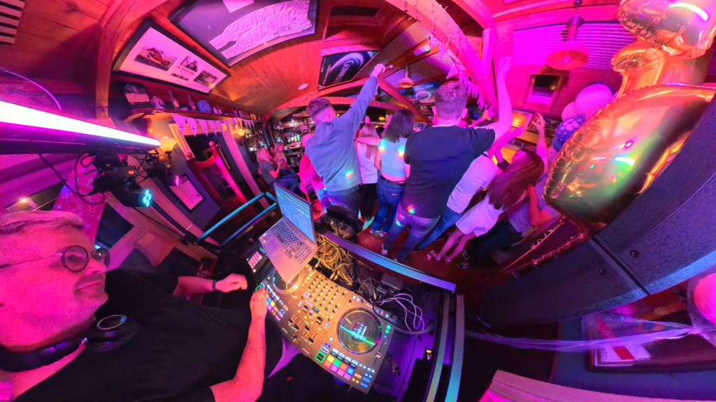 A dynamic, ultra-wide-angle shot from above the DJ's setup, showing DJ William Croke smiling at the crowd reaction. The perspective looks out over a crowded dance floor bathed in vibrant pink and purple lights. The wooden walls are lined with framed pictures.